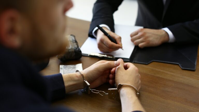 Police officer interrogating criminal in handcuffs at desk indoors